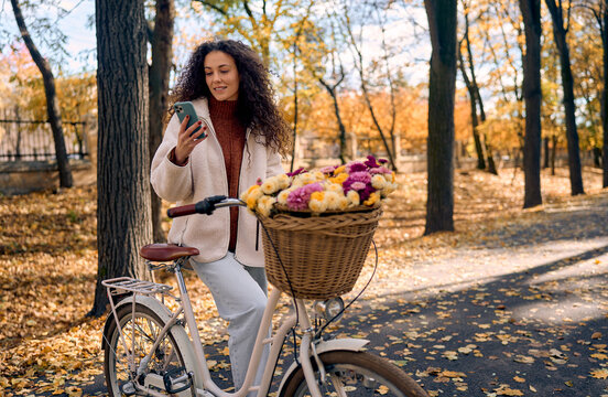 Smiling woman with curly hair using a smartphone while standing next to a white classic city bicycle with a wicker basket full of colorful flowers in a beautiful autumn park with golden fall leaves.