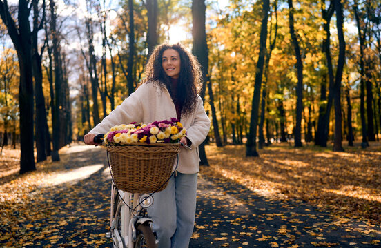 Portrait of beautiful smiling woman with curly hair standing beside a white classic city bicycle with a wicker basket full of colorful flowers in a beautiful autumn park.