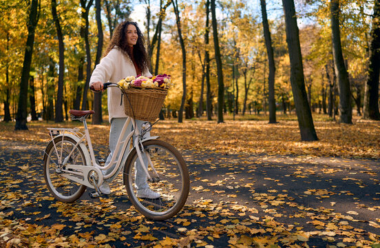 Smiling young woman with curly hair standing beside a white classic city bicycle with a wicker basket full of colorful flowers in a beautiful autumn park with golden fall foliage and warm sunlight
