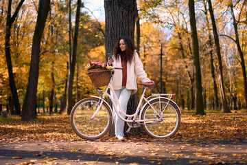 Fototapeta premium Young woman standing in a beautiful autumn park with a white vintage bicycle featuring a wicker basket full of flowers, looking at her smartphone. Enjoying a peaceful autumn day outdoors.