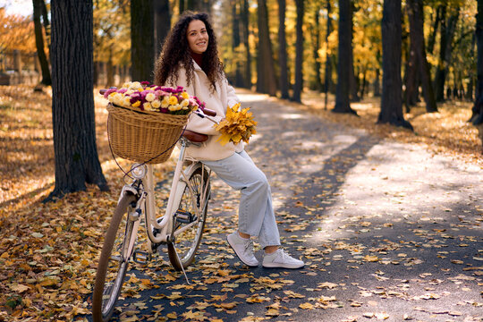 Smiling woman with a flower-filled bicycle pauses on an autumn forest path, surrounded by golden leaves and warm sunlight.