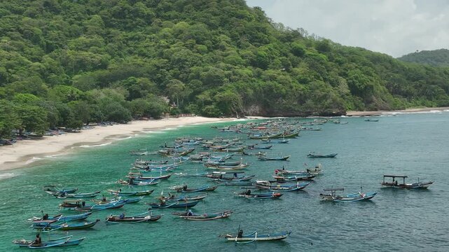 Aerial view of Papuma Beach with numerous traditional fishing boats anchored in the clear turquoise water near the lush green coastline in Jember, East Java, Indonesia.