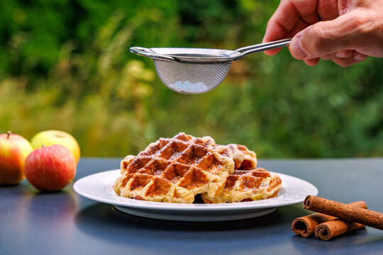 Hand dusting powdered sugar with sieve on homemade Belgian waffles with fresh apples and cinnamon sticks outdoor, autumn dessert concept