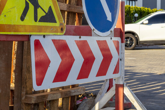 Construction signs show a warning for road work in progress. The signs are positioned near a busy street with cars parked and driving by in the background during daylight hours
