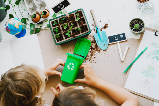 Two young children in white t-shirts sit on the floor and play with a mini green recycling bin. Creative home laboratory concept for ecology and futurism experiments.