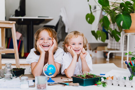Two smiling children wearing white t-shirts sit indoors and pose next to their gardening experiments. Creative home laboratory concept for ecology and futurism experiments.