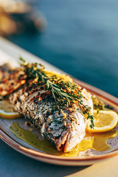 A grilled fish seasoned with fresh herbs (rosemary and thyme), lemon slices, and a drizzle of golden extra virgin olive oil on a ceramic plate.