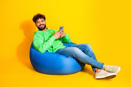 Smiling young man in green hoodie relaxing on blue beanbag while using smartphone against bright yellow background