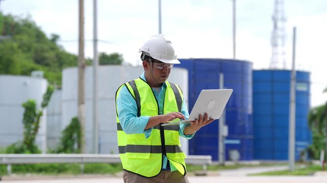 Engineer Oil Industry Worker Wearing Safety Vest and Hard Hat Using Laptop for Industrial Storage Tank Inspection, Maintenance, Technology, and Workplace Safety Outdoors