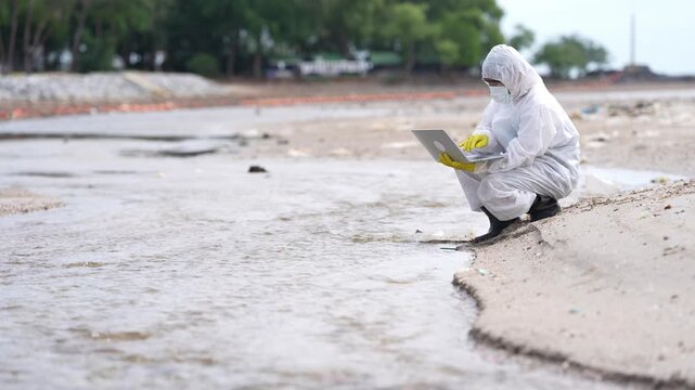 Environmental scientist collecting water sample from floodwater muddy shoreline for pollution research, public health, water testing, environmental monitoring, and fieldwork protective clothing with