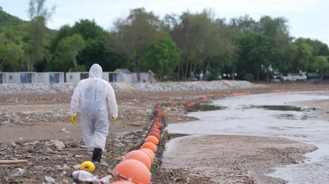 Coastal Cleanup Worker in Protective Suit Inspecting Floating Barrier Along Polluted Shoreline After Low Tide, Supporting Floodwater Environmental Protection and Marine Conservation Efforts