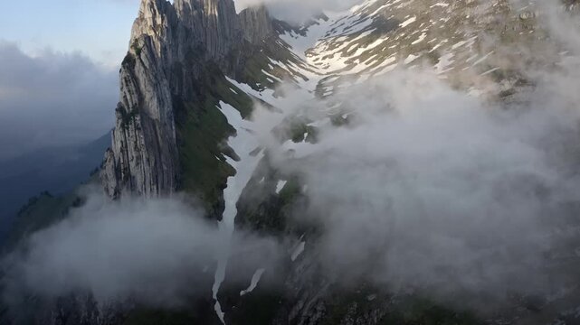 Stunning aerial view of majestic mountain peaks shrouded in ethereal fog and clouds, revealing rugged slopes and patches of snow.