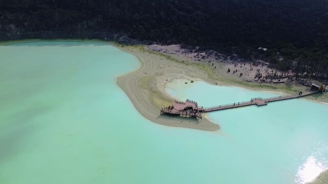 Aerial view of the turquoise Kawah Putih Ciwidey crater lake with a wooden pier and visitors, surrounded by lush forest in Bandung, West Java, Indonesia.