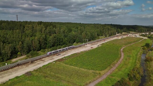 Drone view of a high speed electric train moving parallel to a dense forest with railway tracks crossing a peaceful green landscape during the day