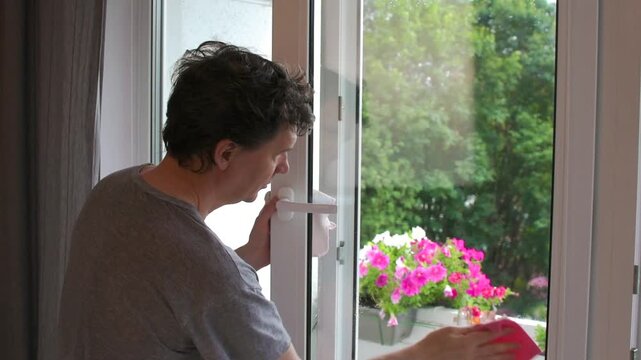 A man cleans the windows in his apartment during his daily routine. He uses a spray and cloth to wipe away dirt and dust from the glass