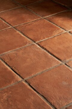 A close view of worn terracotta floor tiles shows varied earthy tones, irregular edges, matte surfaces, and dark grout lines arranged in a simple geometric pattern under warm natural light