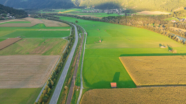 High angle aerial shot displays scenic rural landscape of Lind im Drautal village. Winding Drava river, agricultural fields, main road, and railway line cross lush green valley surrounded by forested 