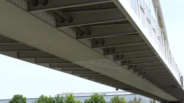 A low-angle, architectural shot looking up at the underside of a modern metal pedestrian or road bridge. The intricate framework of steel girders, beams, and rivets showcases industrial engineering an