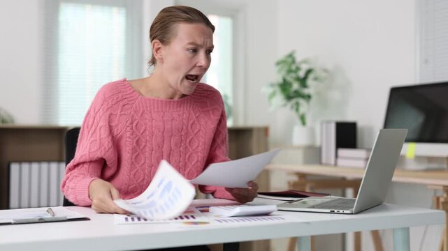 Angry businesswoman shouting during a video call in her office while holding documents, expressing rage, frustration, and disappointment due to work-related stress and problems