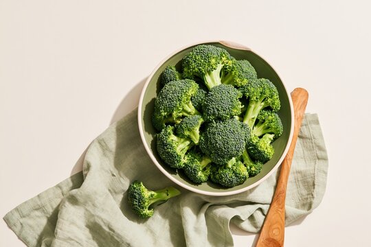 Ceramic bowl filled with fresh broccoli florets on a crumpled cloth with a wooden spoon, featuring organic textures and copy space for plant-based recipes