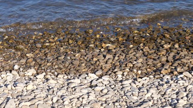 A close-up, high-angle shot of a pebble-covered shoreline next to a flowing river. The transition from dry, light grey stones at the bottom to wet, glistening dark brown stones near the water creates 