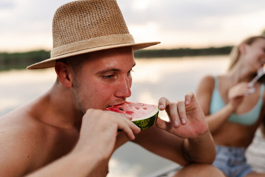 Teen Enjoys Watermelon by River During Summer Afternoon With Friend Nearby