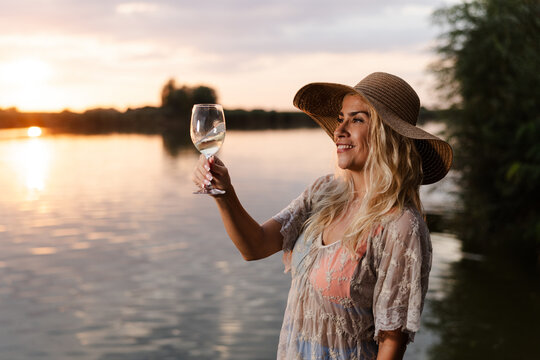 Woman Enjoys Summer by the River While Holding a Glass of Wine During Sunset