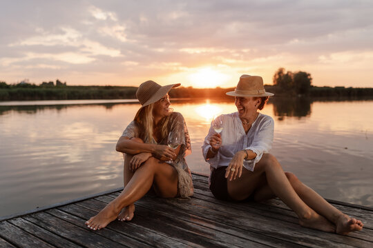 Two Women Enjoy a Summer Evening by the River While Sitting on a Dock and Drinking Wine