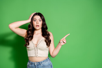 Surprised young woman pointing to the side against a green background wearing a lace-up crop top...