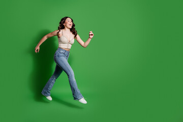 Young woman walking and smiling in casual jeans and crop top against a green studio background,...