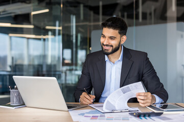 Obraz premium Smiling young businessman reviewing documents and using a laptop at his office desk, symbolizing professional success, financial analysis, and modern business productivity