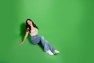 Smiling young woman sitting on green background in casual jeans and white sneakers, relaxed pose...