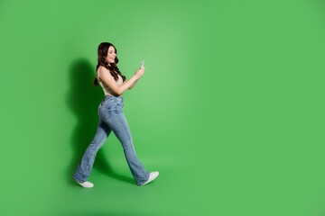 Young woman walking and smiling while looking at smartphone against a green background in casual...