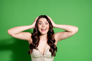 Young excited woman with hands on head smiling in surprise against a green background, joyful...