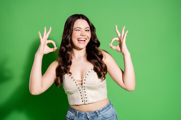 Happy young woman winking and showing OK hand gestures with both hands while smiling against a...