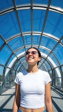 Smiling young woman wearing sunglasses under glass bridge with blue sky and metal structure, expressing happiness during outdoor urban walk
