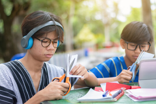 Two Asian teenage boys in traditional clothing focusing on cutting paper with scissors for a school art project at an outdoor green table.