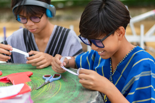 Two happy teenage boys in traditional shirts smiling and holding paper rolls for an art craft project together at an outdoor green table.