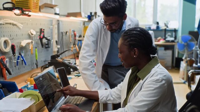 Portrait of African American young adult woman and Indian young adult man in lab coats looking at camera after coding on laptop, camera zooming out. Clip is ideal for STEM education promos