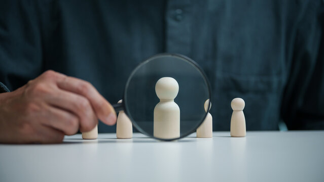 Business professional uses a magnifying glass to focus on a wooden candidate figure, representing recruitment, employee selection, hiring decisions, talent search and workforce management.