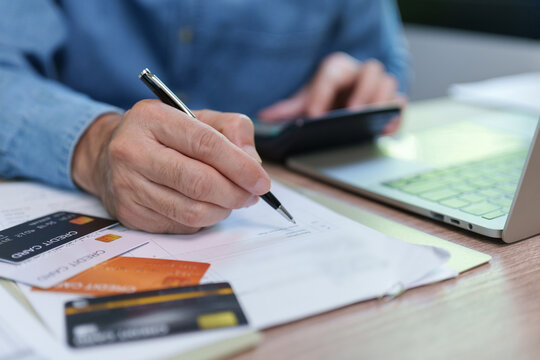 Businessman reviewing financial documents and writing notes beside credit cards, calculator and laptop, representing accounting, debt management, expense reports, budgeting and financial planning.