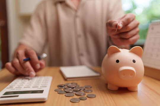 Asian man inserts a coin into a piggy bank beside a calculator and savings, representing personal finance, budgeting, financial planning, investment goals and preparation for the future.