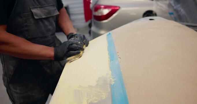 Handheld tracking shot of technician sanding car hood in auto body shop close up on gloved hands smoothing primer along taped seam.