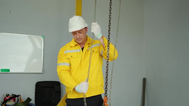 Professional male worker in yellow safety jacket and hard hat pulling a metal chain hoist in a workshop. Manual labor, heavy lifting preparation, safety gear, manufacturing or warehouse concept.