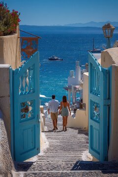 Couple walking through blue gate toward sunlit Aegean Sea, framed by whitewashed coastal village buildings, stone path, bougainvillea, terracotta pots, and bright Mediterranean light