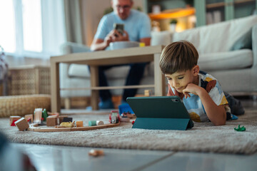 Young boy using tablet on floor while father uses phone © Miljan Živković