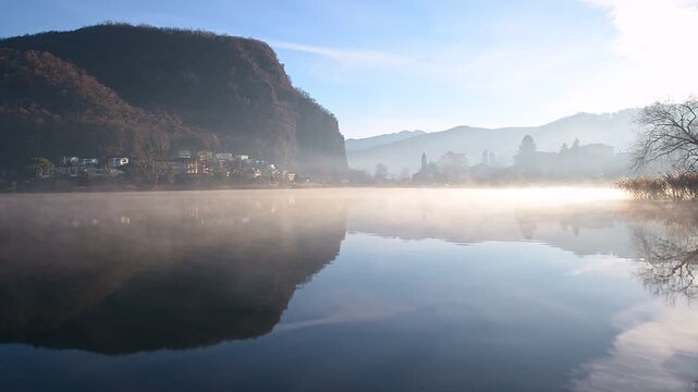 Winter morning fog drifting over Lake Lugano between Switzerland and Italy, calm water and backlit mist at Lavena Strait near Caslano and Lavena Ponte Tresa