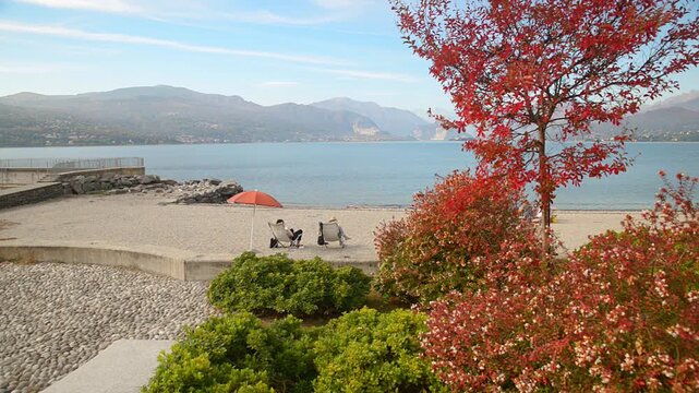 Lake Maggiore beach in Reno with umbrella and deckchairs, calm water, sunny summer day, tranquil lakeside scene in Italy, near hermitage of Santa Caterina del Sasso, Mottarone panoramic view