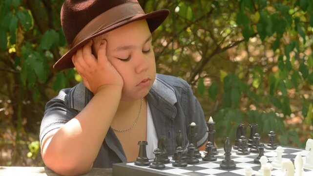 Young Boy Wearing Hat Thinking While Playing Chess Outdoors. A young boy wearing a hat sitting outdoors in a forest, leaning on his hand and thoughtfully considering his next move in a chess game.