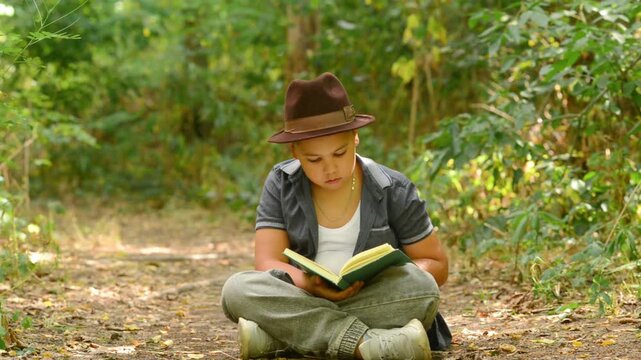 Young Boy Reading Book, Closes It and Dreams Outdoors in Nature Forest. A young boy sitting on the ground in a forest, actively reading a book, then closes it and hugs it tightly. 